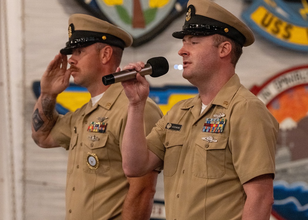 Two U.S. Navy enlisted personnel in khaki service uniforms are participating in a formal military ceremony. The sailor in the foreground is speaking into a microphone, while the sailor behind him renders a salute. The setting appears to be aboard a naval vessel, indicated by the 'USS' insignia and ship crests visible on the wall behind them.