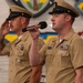Two U.S. Navy enlisted personnel in khaki service uniforms are participating in a formal military ceremony. The sailor in the foreground is speaking into a microphone, while the sailor behind him renders a salute. The setting appears to be aboard a naval vessel, indicated by the 'USS' insignia and ship crests visible on the wall behind them.