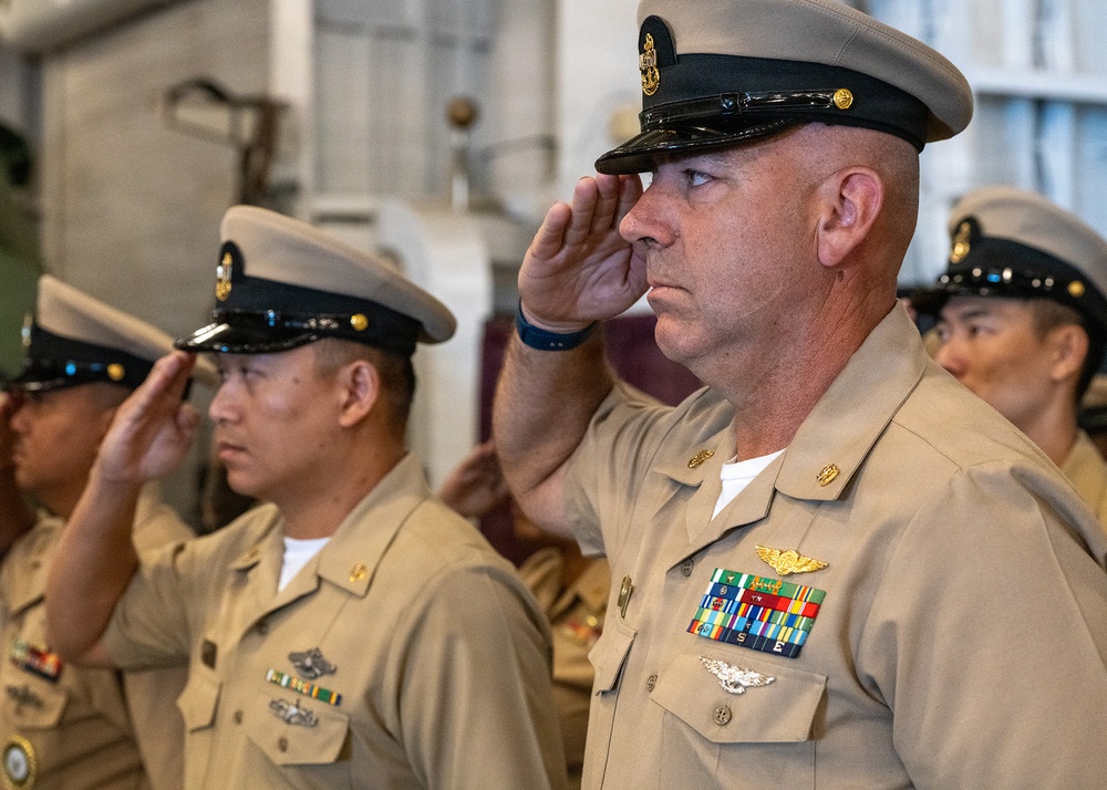 Multiple U.S. Navy enlisted personnel in Service Khaki (Type B) uniforms are rendering a salute during a formal military ceremony. The foreground subject displays aviation warfare insignia (Naval Aviator wings), multiple service ribbons, and a Master Chief Petty Officer cap device. The setting appears to be an indoor shipboard or hangar bay environment.