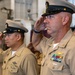Multiple U.S. Navy enlisted personnel in Service Khaki (Type B) uniforms are rendering a salute during a formal military ceremony. The foreground subject displays aviation warfare insignia (Naval Aviator wings), multiple service ribbons, and a Master Chief Petty Officer cap device. The setting appears to be an indoor shipboard or hangar bay environment.