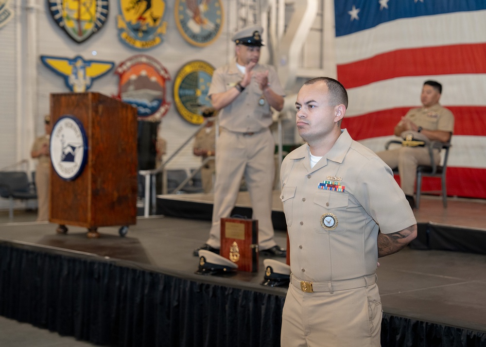 A formal U.S. Navy military ceremony is taking place in what appears to be a hangar or large indoor facility. A Navy enlisted sailor in khaki service uniform stands at attention in the foreground, while an officer speaks at a podium on a raised stage behind him. Multiple military unit insignia/seals are displayed on the wall, and a large American flag is prominently visible in the background.