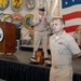 A formal U.S. Navy military ceremony is taking place in what appears to be a hangar or large indoor facility. A Navy enlisted sailor in khaki service uniform stands at attention in the foreground, while an officer speaks at a podium on a raised stage behind him. Multiple military unit insignia/seals are displayed on the wall, and a large American flag is prominently visible in the background.