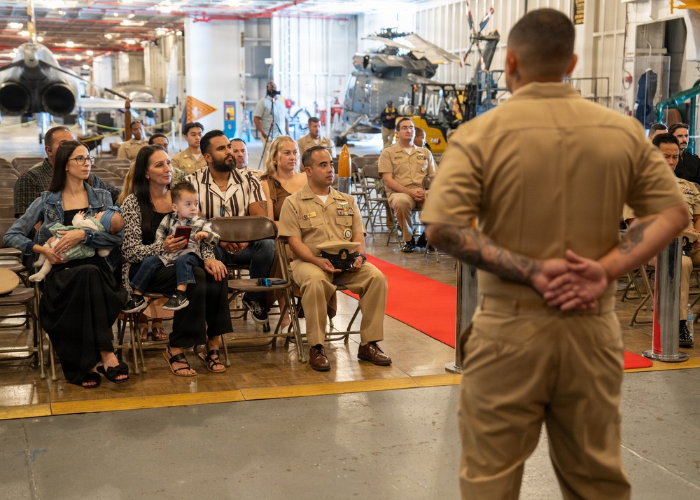 A military ceremony is taking place inside what appears to be a naval hangar bay, with aircraft and helicopters visible in the background. A uniformed service member stands with hands clasped behind their back, addressing a seated audience that includes both military personnel in tan/khaki uniforms and civilian family members. A red carpet runner is visible on the floor, suggesting a formal occasion such as a promotion, retirement, or re-enlistment ceremony.
