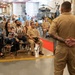 A military ceremony is taking place inside what appears to be a naval hangar bay, with aircraft and helicopters visible in the background. A uniformed service member stands with hands clasped behind their back, addressing a seated audience that includes both military personnel in tan/khaki uniforms and civilian family members. A red carpet runner is visible on the floor, suggesting a formal occasion such as a promotion, retirement, or re-enlistment ceremony.