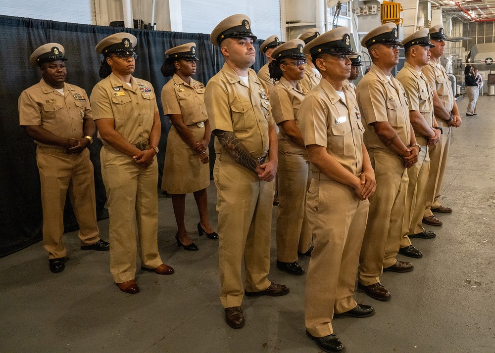 A group of U.S. Navy personnel in khaki service uniforms are standing at attention in a formal formation, likely during a ceremony or official event. They are positioned in a row inside what appears to be a large hangar bay or industrial military facility. The personnel display a mix of ranks and genders, all wearing the standard Navy Chief Petty Officer or senior enlisted khaki uniform.