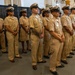 A group of U.S. Navy personnel in khaki service uniforms are standing at attention in a formal formation, likely during a ceremony or official event. They are positioned in a row inside what appears to be a large hangar bay or industrial military facility. The personnel display a mix of ranks and genders, all wearing the standard Navy Chief Petty Officer or senior enlisted khaki uniform.