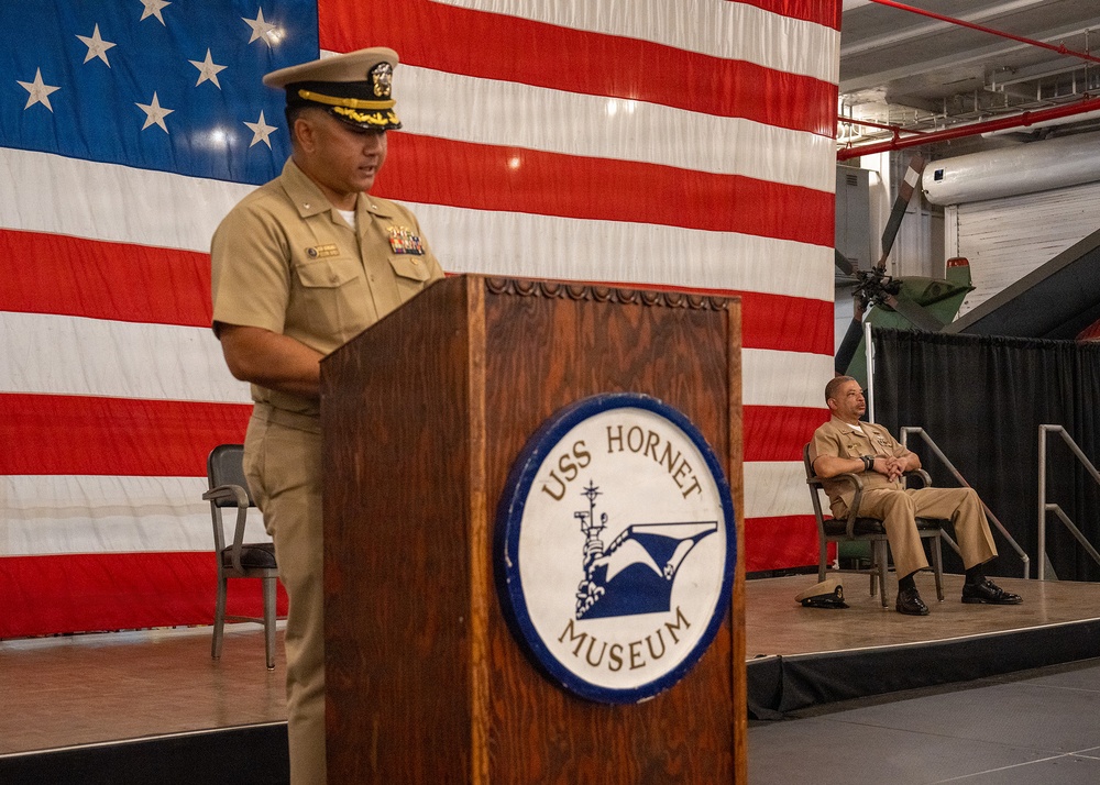 A U.S. Navy officer in service khaki uniform is speaking at a podium bearing the USS Hornet Museum seal, with a large American flag draped as a backdrop. A second Navy officer is seated on the stage behind the speaker. The setting appears to be an official military ceremony held inside the USS Hornet Museum's hangar deck.