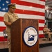 A U.S. Navy officer in service khaki uniform is speaking at a podium bearing the USS Hornet Museum seal, with a large American flag draped as a backdrop. A second Navy officer is seated on the stage behind the speaker. The setting appears to be an official military ceremony held inside the USS Hornet Museum's hangar deck.