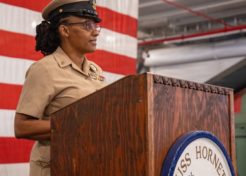 A U.S. Navy enlisted service member in khaki uniform stands at a wooden podium bearing the 'USS Hornet' insignia, apparently delivering a speech or address. An American flag is visible in the background. The setting appears to be inside a hangar bay or large interior space aboard or associated with the USS Hornet.