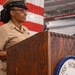 A U.S. Navy enlisted service member in khaki uniform stands at a wooden podium bearing the 'USS Hornet' insignia, apparently delivering a speech or address. An American flag is visible in the background. The setting appears to be inside a hangar bay or large interior space aboard or associated with the USS Hornet.