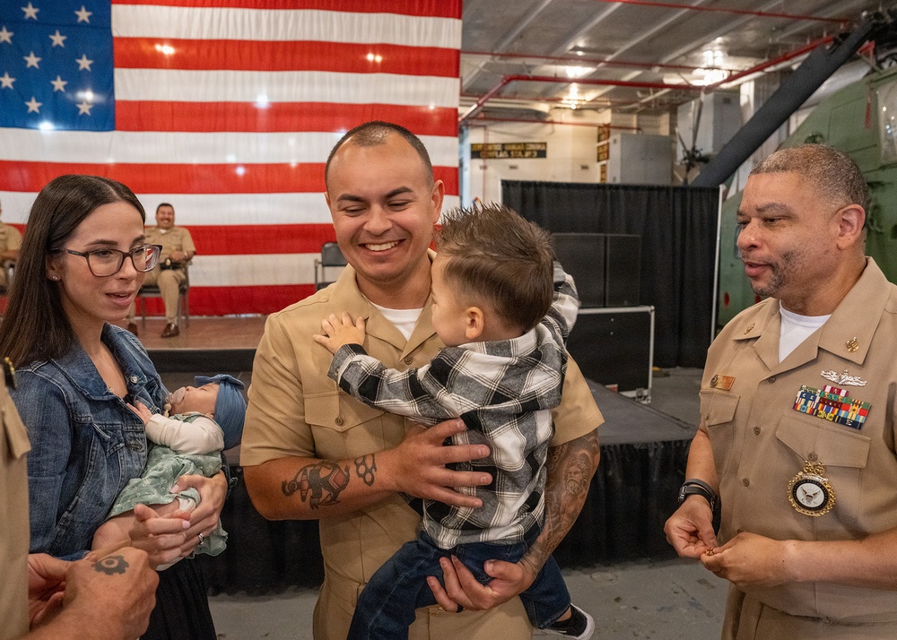 A U.S. Navy service member in tan/khaki uniform joyfully holds a toddler during what appears to be a military ceremony or homecoming event. A woman holding an infant stands beside him, while a senior Navy officer with multiple ribbons and medals observes nearby. A large American flag is displayed on a stage backdrop in the background.