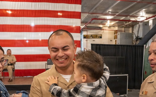 A U.S. Navy service member in tan/khaki uniform joyfully holds a toddler during what appears to be a military ceremony or homecoming event. A woman holding an infant stands beside him, while a senior Navy officer with multiple ribbons and medals observes nearby. A large American flag is displayed on a stage backdrop in the background.
