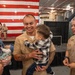 A U.S. Navy service member in tan/khaki uniform joyfully holds a toddler during what appears to be a military ceremony or homecoming event. A woman holding an infant stands beside him, while a senior Navy officer with multiple ribbons and medals observes nearby. A large American flag is displayed on a stage backdrop in the background.
