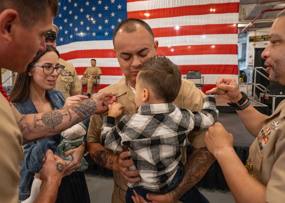 A U.S. Marine in dress tan uniform is holding a young child during what appears to be a military promotion or pinning ceremony. Family members and fellow service members are pinning new rank insignia onto the Marine's collar while he holds the child. The event is taking place in front of a large American flag, consistent with a formal military ceremony setting.