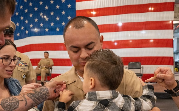A U.S. Marine in dress tan uniform is holding a young child during what appears to be a military promotion or pinning ceremony. Family members and fellow service members are pinning new rank insignia onto the Marine's collar while he holds the child. The event is taking place in front of a large American flag, consistent with a formal military ceremony setting.