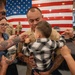 A U.S. Marine in dress tan uniform is holding a young child during what appears to be a military promotion or pinning ceremony. Family members and fellow service members are pinning new rank insignia onto the Marine's collar while he holds the child. The event is taking place in front of a large American flag, consistent with a formal military ceremony setting.