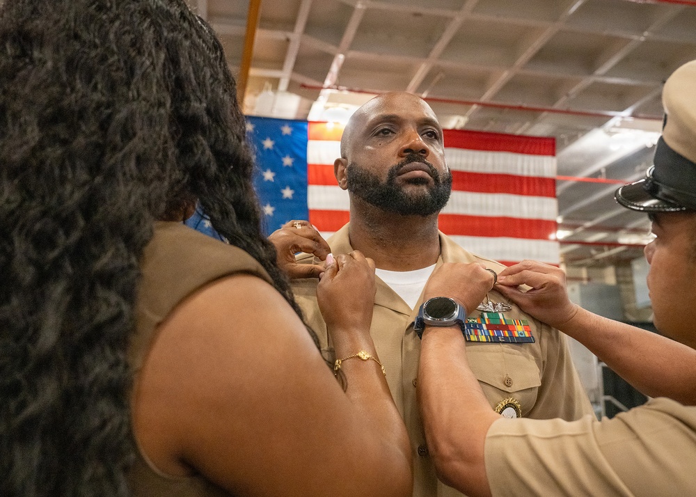 A U.S. Navy service member is having insignia or rank devices pinned onto the collar of his khaki uniform during what appears to be a promotion or advancement ceremony. Two individuals — one in civilian attire on the left and one in Navy uniform on the right — are simultaneously pinning devices to each side of his collar. An American flag is displayed prominently in the background.