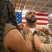 A U.S. Navy service member is having insignia or rank devices pinned onto the collar of his khaki uniform during what appears to be a promotion or advancement ceremony. Two individuals — one in civilian attire on the left and one in Navy uniform on the right — are simultaneously pinning devices to each side of his collar. An American flag is displayed prominently in the background.