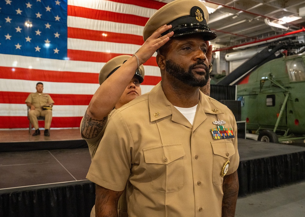 A U.S. Navy Chief Petty Officer is having his combination cover (hat) placed on his head during what appears to be a Chief Petty Officer pinning or frocking ceremony. The event is taking place in what appears to be a hangar bay or museum setting with a large American flag as a backdrop. A military helicopter and other personnel in Navy khaki uniforms are visible in the background.