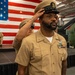A U.S. Navy Chief Petty Officer is having his combination cover (hat) placed on his head during what appears to be a Chief Petty Officer pinning or frocking ceremony. The event is taking place in what appears to be a hangar bay or museum setting with a large American flag as a backdrop. A military helicopter and other personnel in Navy khaki uniforms are visible in the background.