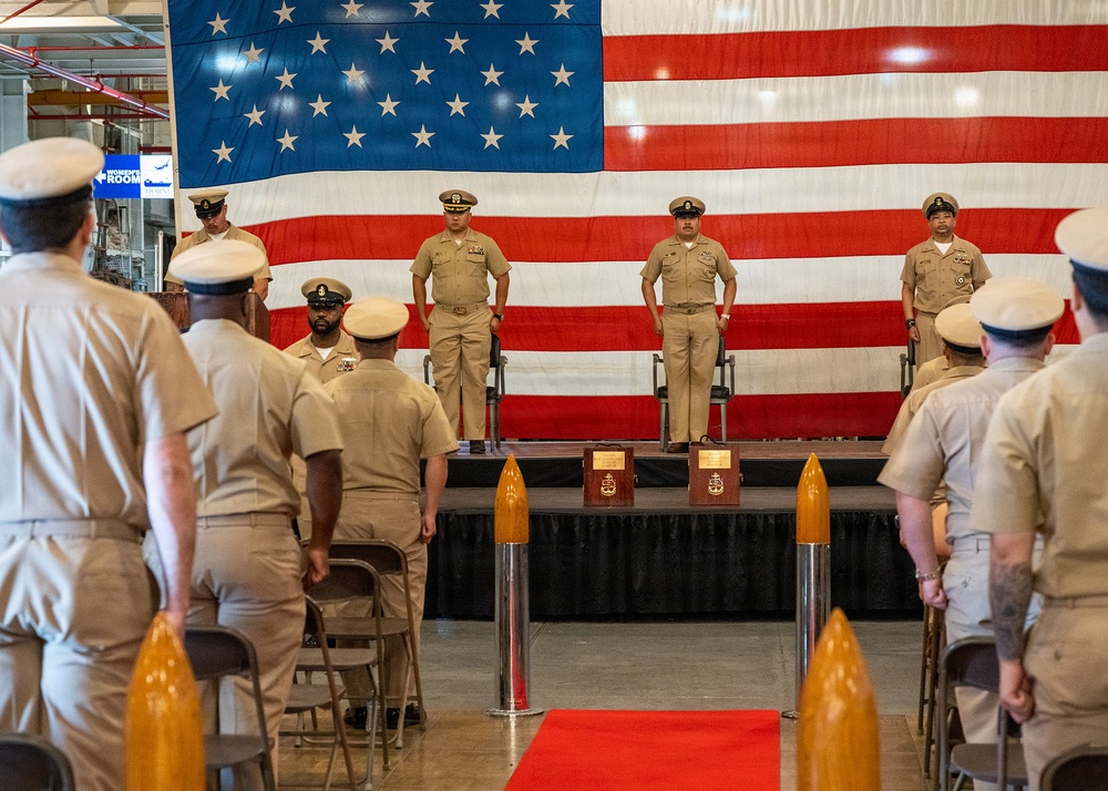 A formal U.S. Navy military ceremony is taking place indoors, with personnel in khaki service uniforms standing at attention on and before a raised stage. A large American flag serves as the backdrop, and decorative torpedo-shaped bollards line a red carpet aisle. Two plaques on wooden bases are displayed on the stage, suggesting a change of command or award ceremony.