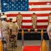 A formal U.S. Navy military ceremony is taking place indoors, with personnel in khaki service uniforms standing at attention on and before a raised stage. A large American flag serves as the backdrop, and decorative torpedo-shaped bollards line a red carpet aisle. Two plaques on wooden bases are displayed on the stage, suggesting a change of command or award ceremony.