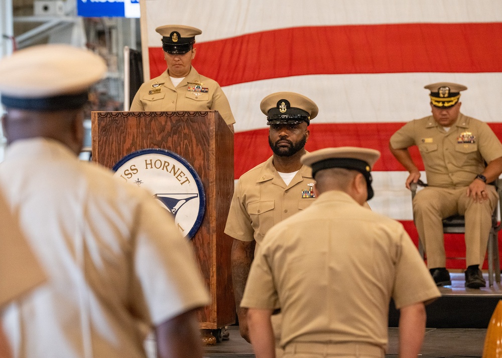 A formal U.S. Navy ceremony is taking place aboard or at a venue associated with the USS Hornet, as indicated by the podium seal. Several Navy personnel in tan/khaki service uniforms are present on and around a stage, with a large American flag displayed in the background. The scene appears to be a change of command, promotion, or chief petty officer pinning ceremony.