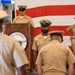 A formal U.S. Navy ceremony is taking place aboard or at a venue associated with the USS Hornet, as indicated by the podium seal. Several Navy personnel in tan/khaki service uniforms are present on and around a stage, with a large American flag displayed in the background. The scene appears to be a change of command, promotion, or chief petty officer pinning ceremony.