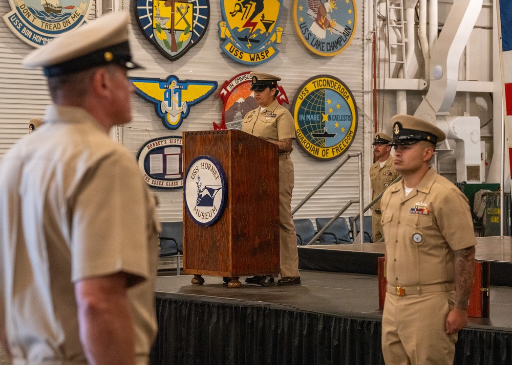 A U.S. Navy ceremony is taking place aboard or inside the USS Hornet Museum, with a uniformed sailor speaking at a podium bearing the USS Hornet Museum seal. Multiple Navy personnel in tan/khaki service uniforms are present, some standing at attention. The backdrop features numerous naval unit insignia patches and plaques from various USS vessels.