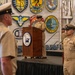 A U.S. Navy ceremony is taking place aboard or inside the USS Hornet Museum, with a uniformed sailor speaking at a podium bearing the USS Hornet Museum seal. Multiple Navy personnel in tan/khaki service uniforms are present, some standing at attention. The backdrop features numerous naval unit insignia patches and plaques from various USS vessels.