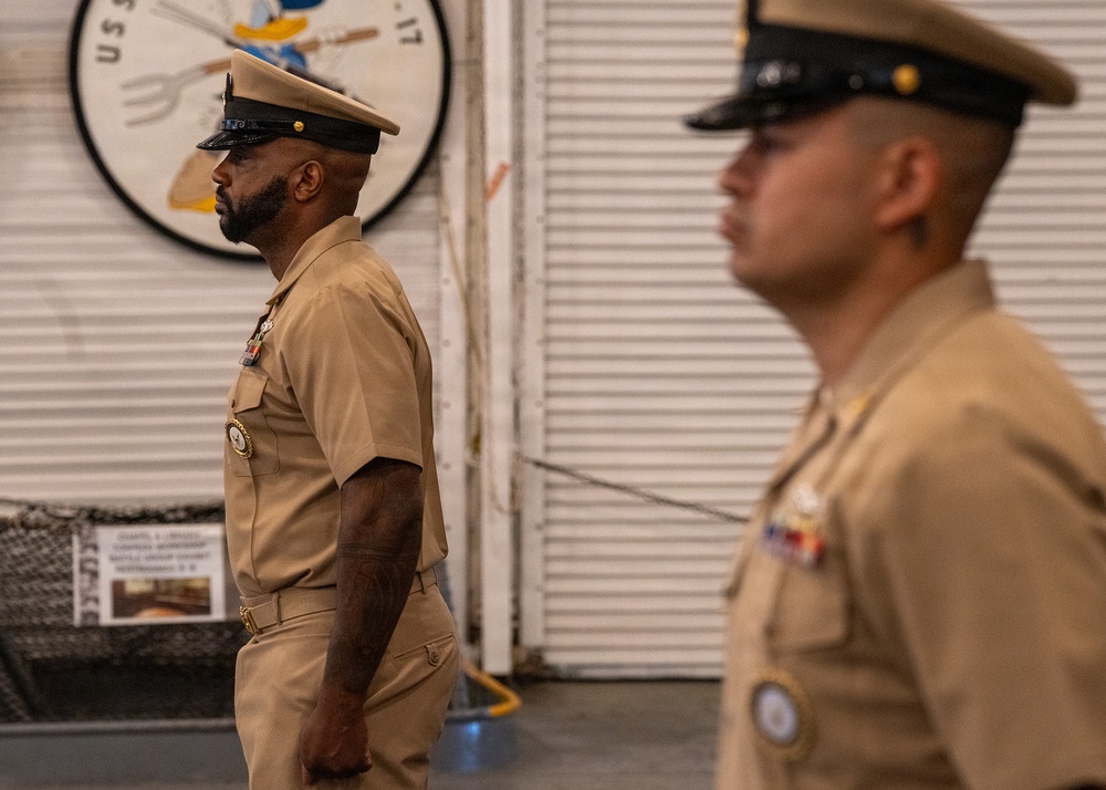 Two U.S. Navy personnel in service khaki uniforms are standing at attention or in a formal posture inside what appears to be a naval vessel hangar bay. Both wear combination covers (officer/chief petty officer caps) and display ribbons and insignia on their uniforms. A circular unit insignia patch featuring a Donald Duck-like cartoon character is visible on the wall behind them.