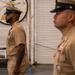 Two U.S. Navy personnel in service khaki uniforms are standing at attention or in a formal posture inside what appears to be a naval vessel hangar bay. Both wear combination covers (officer/chief petty officer caps) and display ribbons and insignia on their uniforms. A circular unit insignia patch featuring a Donald Duck-like cartoon character is visible on the wall behind them.