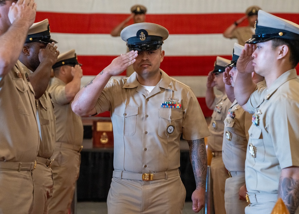 A U.S. Navy ceremony is taking place in front of a large American flag, with multiple sailors in khaki service uniforms rendering salutes. The central figure, a Chief Petty Officer identifiable by his combination cover with anchor insignia and multiple service ribbons and medals, is returning a salute while walking through a corridor formed by fellow sailors. The formal nature of the event, the decorations, and the lined formation suggest this is a pinning ceremony, promotion ceremony, or similar official military event.