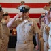 A U.S. Navy ceremony is taking place in front of a large American flag, with multiple sailors in khaki service uniforms rendering salutes. The central figure, a Chief Petty Officer identifiable by his combination cover with anchor insignia and multiple service ribbons and medals, is returning a salute while walking through a corridor formed by fellow sailors. The formal nature of the event, the decorations, and the lined formation suggest this is a pinning ceremony, promotion ceremony, or similar official military event.