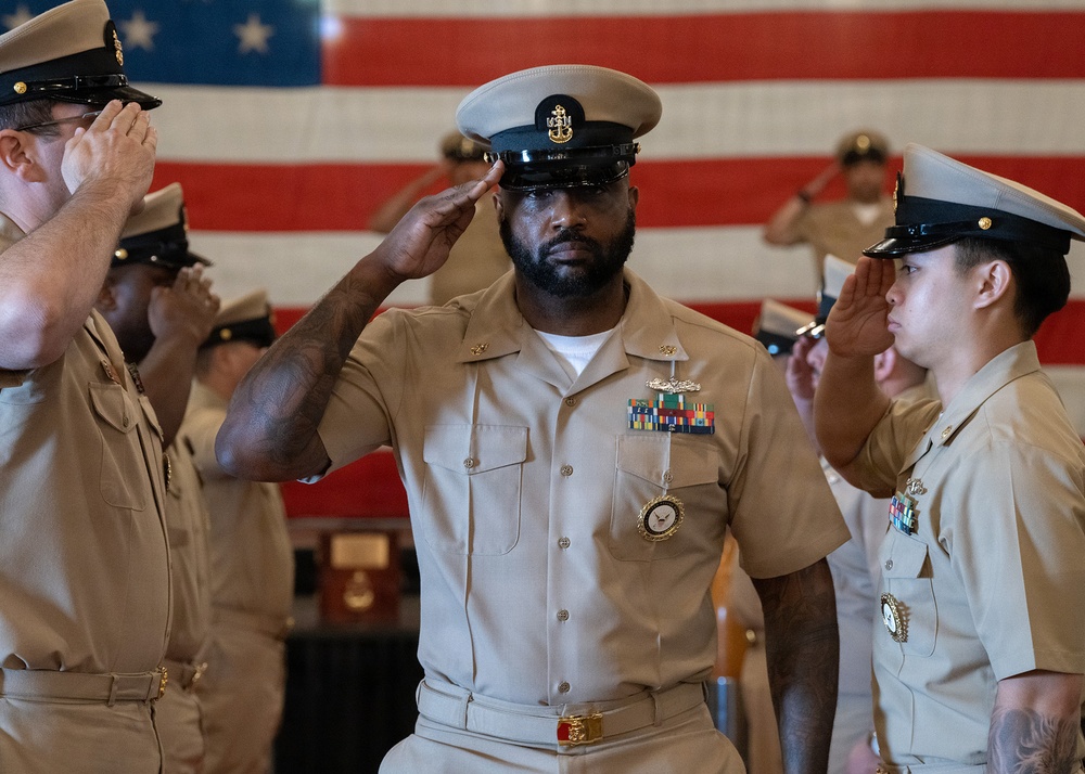 A group of U.S. Navy personnel in Service Khaki (Type I) uniforms are rendering salutes during a formal military ceremony. The central figure, a senior enlisted sailor wearing a combination cover with a Master Chief or Senior Chief insignia and displaying multiple service ribbons and a large medallion, appears to be the focal point of the event. A large American flag is prominently displayed in the background.