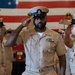 A group of U.S. Navy personnel in Service Khaki (Type I) uniforms are rendering salutes during a formal military ceremony. The central figure, a senior enlisted sailor wearing a combination cover with a Master Chief or Senior Chief insignia and displaying multiple service ribbons and a large medallion, appears to be the focal point of the event. A large American flag is prominently displayed in the background.