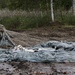 A soldier in camouflage uniform and helmet is kneeling on muddy ground, gathering and organizing parachute suspension lines after a landing. A large gray parachute canopy is spread across the wet, muddy terrain in front of them. The setting appears to be a drop zone near a treeline, likely during a military airborne exercise.