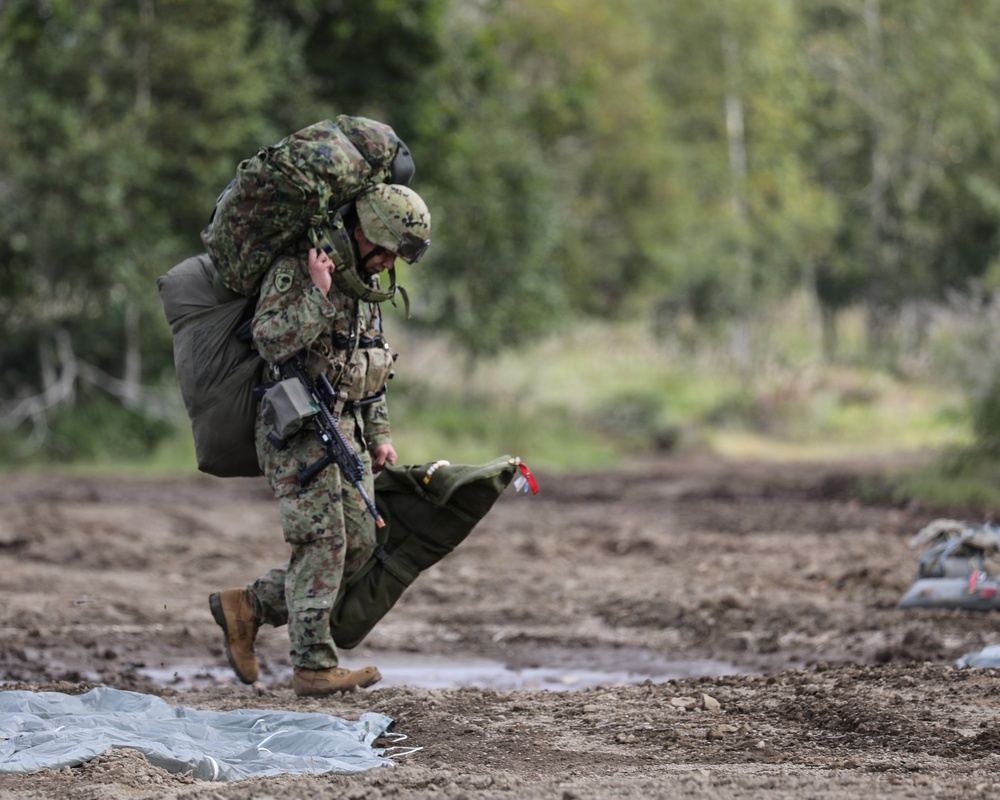A heavily loaded soldier in camouflage uniform is walking across a muddy dirt field, carrying an extremely large pack on their back along with additional gear. The soldier is also holding what appears to be a parachute bag or equipment bag in one hand and has a rifle slung on their body. The setting appears to be a military training drop zone or landing area, with parachute material visible on the ground.
