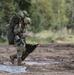 A heavily loaded soldier in camouflage uniform is walking across a muddy dirt field, carrying an extremely large pack on their back along with additional gear. The soldier is also holding what appears to be a parachute bag or equipment bag in one hand and has a rifle slung on their body. The setting appears to be a military training drop zone or landing area, with parachute material visible on the ground.
