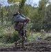 A heavily equipped soldier in full combat gear is moving across a muddy landing zone after an airborne operation. The soldier is carrying a large rucksack with a bundled parachute on top, along with a rifle, suggesting a recent parachute landing. A green parachute canopy is visible in the upper left background, indicating another jumper is still descending.