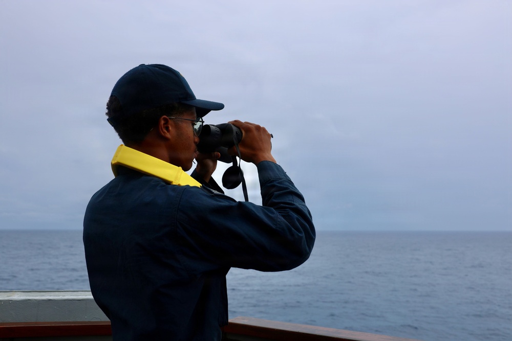 A person wearing a dark navy jacket and cap is standing on the deck of a vessel, looking through binoculars toward the open sea. They are wearing a yellow high-visibility collar or life vest underneath their jacket. The sky is overcast and the ocean appears calm, suggesting maritime patrol or observation duties.