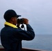 A person wearing a dark navy jacket and cap is standing on the deck of a vessel, looking through binoculars toward the open sea. They are wearing a yellow high-visibility collar or life vest underneath their jacket. The sky is overcast and the ocean appears calm, suggesting maritime patrol or observation duties.