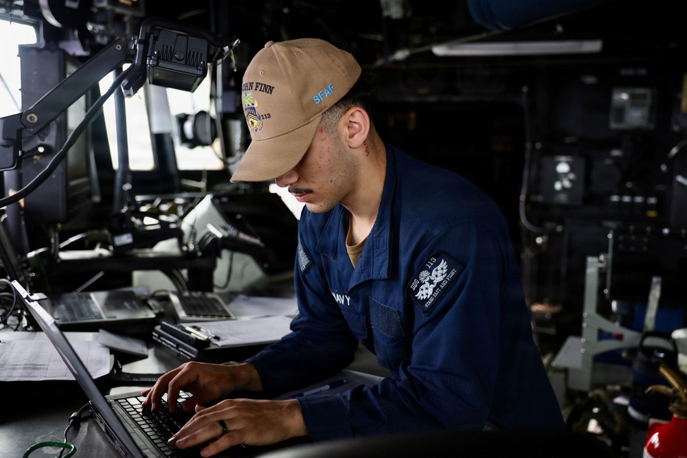 A U.S. Navy sailor is working on a laptop computer aboard what appears to be the bridge or a control station of a naval vessel. The sailor is wearing a blue Navy uniform and a tan cap bearing the name 'John Finn' and the designation 'SFAF', indicating assignment to USS John Finn (DDG-113). The surrounding environment is filled with naval equipment, monitors, and instrumentation typical of a warship's operational spaces.