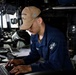 A U.S. Navy sailor is working on a laptop computer aboard what appears to be the bridge or a control station of a naval vessel. The sailor is wearing a blue Navy uniform and a tan cap bearing the name 'John Finn' and the designation 'SFAF', indicating assignment to USS John Finn (DDG-113). The surrounding environment is filled with naval equipment, monitors, and instrumentation typical of a warship's operational spaces.