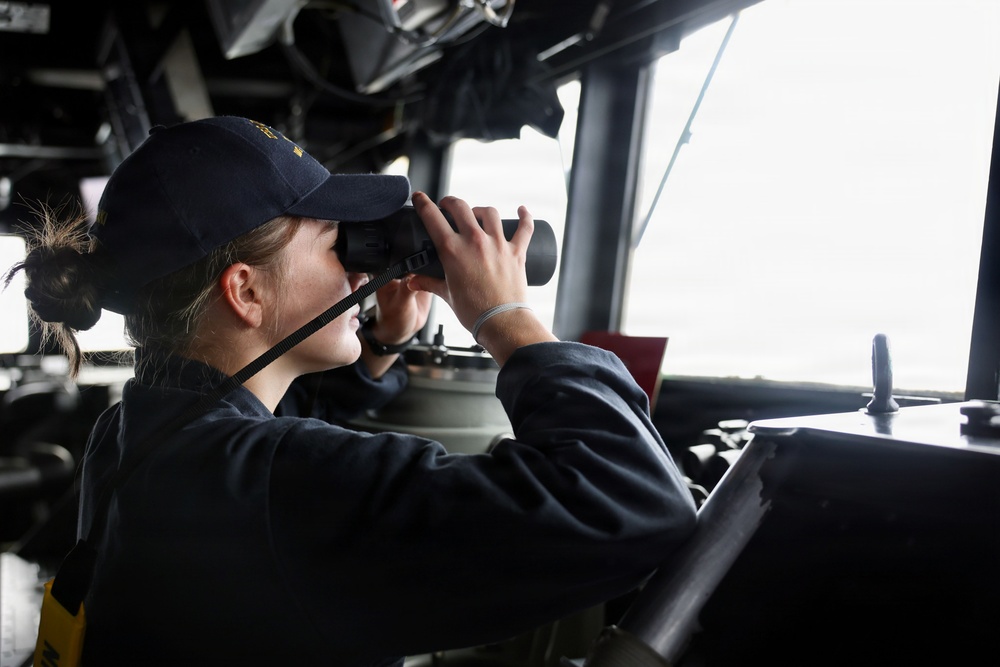 A person wearing a dark navy uniform and cap is using binoculars while stationed at what appears to be the bridge of a naval vessel. They are looking out through large windows toward the horizon. The setting is consistent with a military ship's bridge or command station.