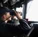 A person wearing a dark navy uniform and cap is using binoculars while stationed at what appears to be the bridge of a naval vessel. They are looking out through large windows toward the horizon. The setting is consistent with a military ship's bridge or command station.