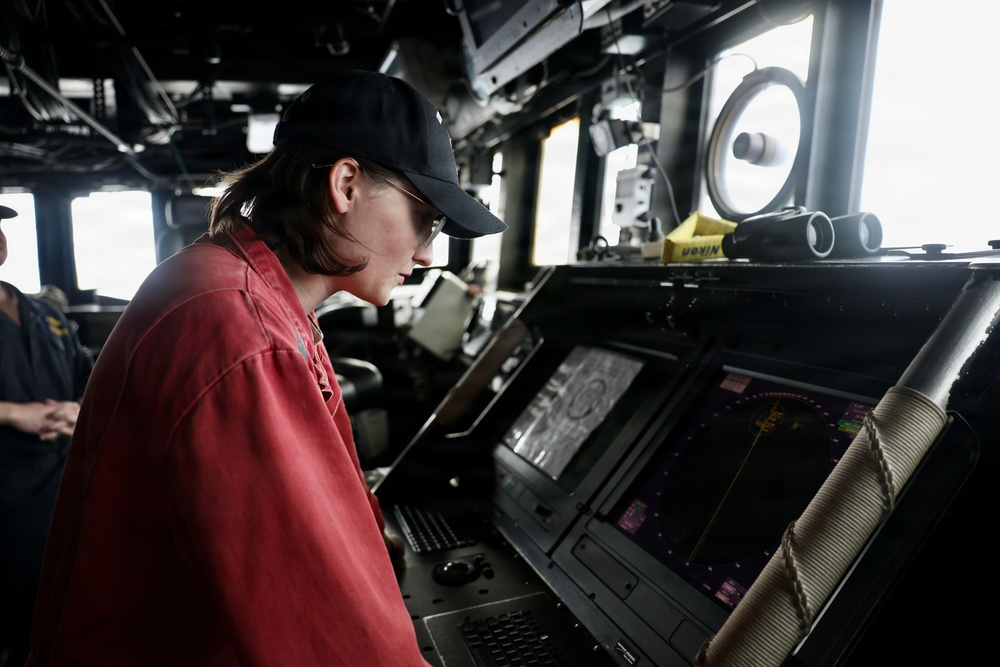 A person wearing a red shirt and black baseball cap is intently studying navigation displays on what appears to be the bridge of a naval vessel. Multiple electronic screens showing radar and navigation data are visible at the console. The setting is a ship's bridge with various instruments, binoculars, and communication equipment visible in the background.