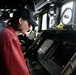 A person wearing a red shirt and black baseball cap is intently studying navigation displays on what appears to be the bridge of a naval vessel. Multiple electronic screens showing radar and navigation data are visible at the console. The setting is a ship's bridge with various instruments, binoculars, and communication equipment visible in the background.