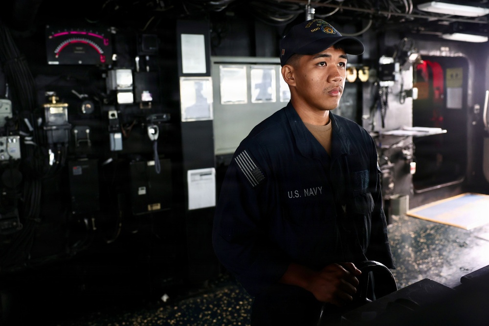 A U.S. Navy sailor stands at a station aboard what appears to be the bridge or combat information center of a naval vessel. The sailor is wearing standard Navy working uniform (NWU) with a U.S. flag patch on the sleeve and a Navy ball cap with a ship's insignia. The environment is dimly lit with various control panels, instruments, and equipment visible in the background.
