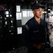 A U.S. Navy sailor stands at a station aboard what appears to be the bridge or combat information center of a naval vessel. The sailor is wearing standard Navy working uniform (NWU) with a U.S. flag patch on the sleeve and a Navy ball cap with a ship's insignia. The environment is dimly lit with various control panels, instruments, and equipment visible in the background.